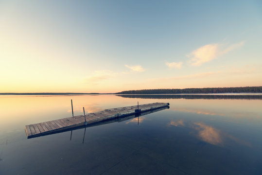 A wooden dock floating on flat calm waters of a lake at sunset.