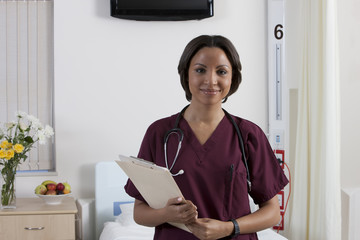 Portrait of smiling medical professional in maroon scrubs standing in a hospital room holding a clipboard