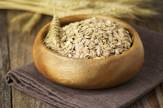 Close-up of organic rolled oats in a wooden bowl on a folded napkin with ears of wheat on a table