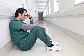 Tired doctor sitting on the floor in a hospital corridor with his hand on his head