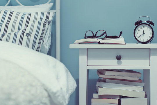 Books and an alarm clock on a bedside table next to a bed in a bedroom