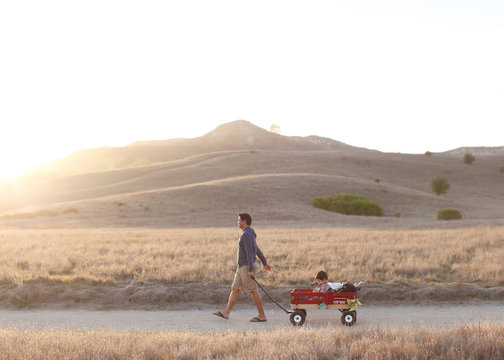 Side view of father pulling son in cart while walking on dirt road