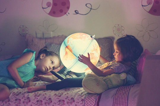 Girls looking at illuminated lamp in shape of globe while lying on bed in bedroom