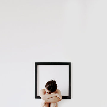 Naked woman sitting in front of a blank picture frame hugging her knees