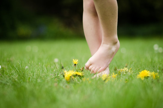 Close-up of a barefoot boy standing on one leg on a lawn with wild dandelions growing in a summer garden