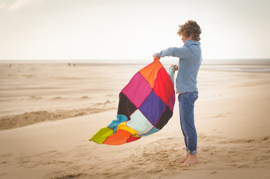 Side view of a barefoot boy in jeans standing on a sandy beach shaking a multi coloured patchwork blanket