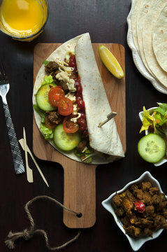 Overhead view of a Shawarma wrap with spiced meat, tomatoes, cucumber, and garlic sauce on a wooden chopping board with a glass of orange juice