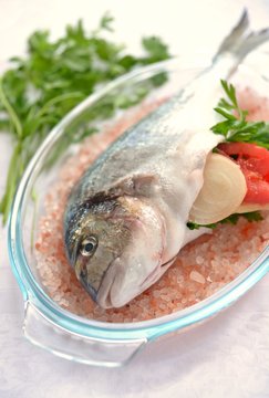 Whole uncooked gilt-head sea bream stuffed with onion, tomato and parsley on a bed of Himalayan salt in a glass dish