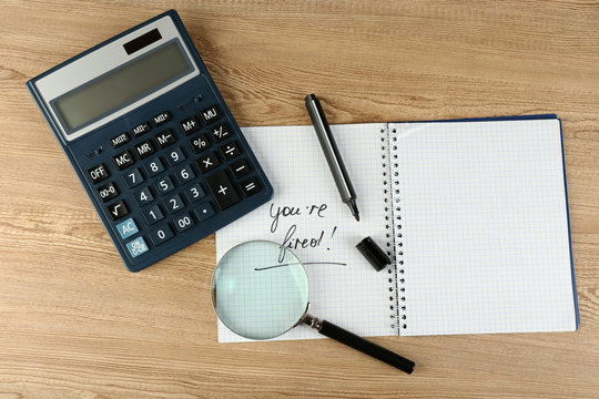 Message You're Fired on notebook with calculator, marker and magnifying glass on wooden table, closeup