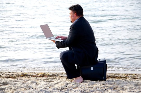 Geschäftsmann sitzt auf seiner Aktentasche am Strand und tippt auf seinem Laptop.