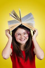 miling Portrait of a cute little schoolgirl loving to learn, holding with hands a book and wearing glasses isolated over yellow background. 