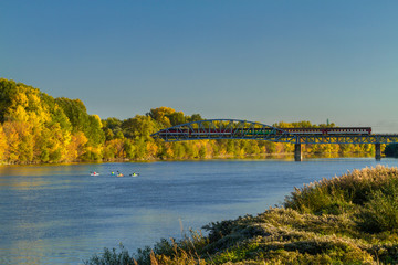 Obraz premium Autumn at river with kayaks and train crossing bridge.