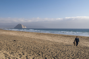 Man and his dog walking on empty beach with Morro Rock on Backgr