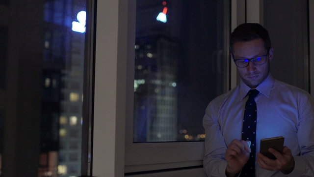Businessman standing next to the window and using smartphone at night
