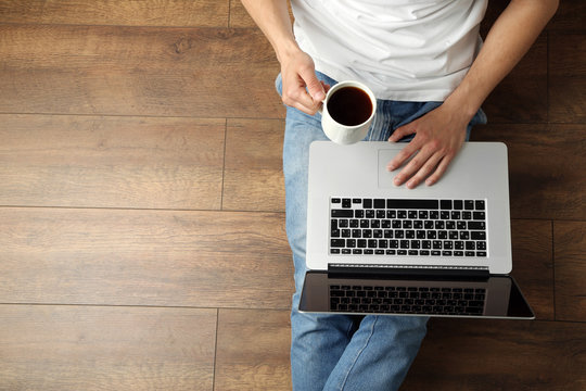 Young Man Sitting On Floor With Laptop And Cup Of Coffee In Room