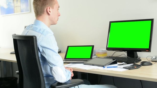 young handsome man sits and looks on desktop and laptop computer in the office - green screen