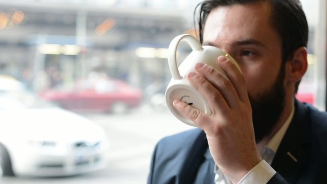 young handsome man with full-beard (hipster) drinks coffee in cafe - urban street in background