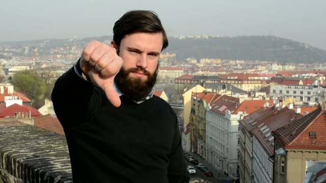 young handsome man with full-beard (hipster) shows thumb on rejection - city in background