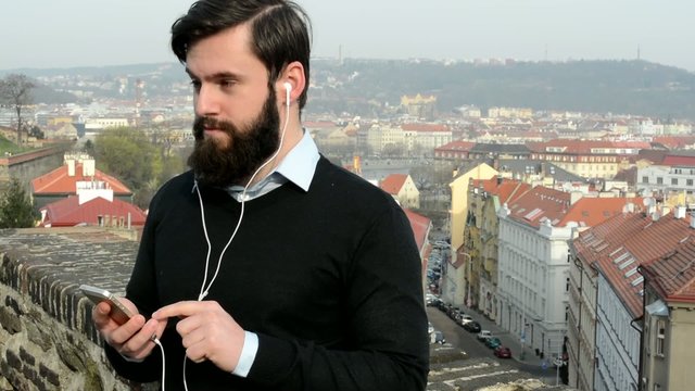 young handsome man with full-beard (hipster) listen music with smartphone - city in background