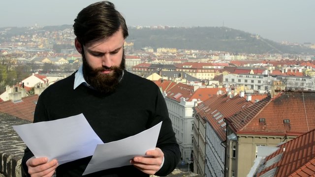 young handsome man with full-beard (hipster) reads documents - city in background