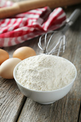 Fototapeta na wymiar Bowl of wheat flour with eggs and whisk on grey wooden backgroun