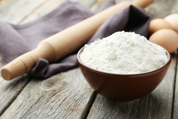 Bowl of wheat flour with eggs and rolling pin on grey wooden bac