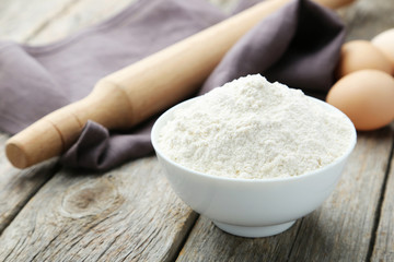 Bowl of wheat flour with eggs and rolling pin on grey wooden bac