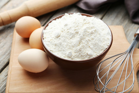 Bowl of wheat flour with eggs and whisk on grey wooden backgroun
