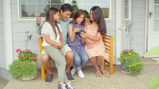 A family sits on a bench outside of their house and uses a tablet