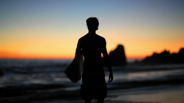 A surfer walks along the beach at sunset checking out the surf
