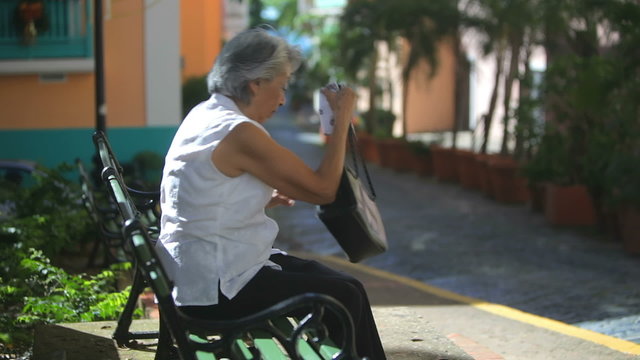 An old lady walks up to a bench, sits down and takes a tablet out of her bag 