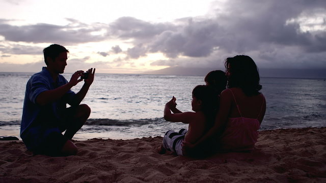 A man taking a picture of his family sitting in the sand at the beach at sunset