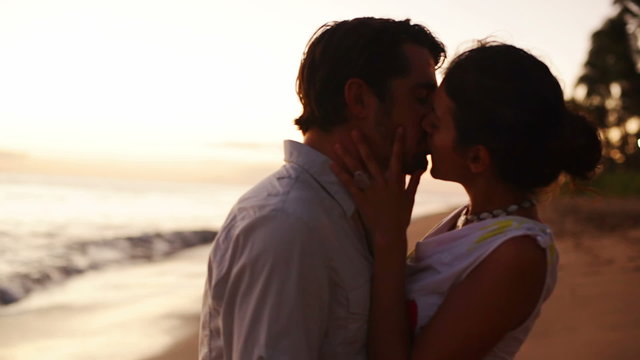 A silhouetted couple hold each other and kiss on the beach during magic hour