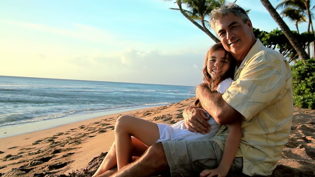 A little girl sitting in her father's lap on the beach in Hawaii.
