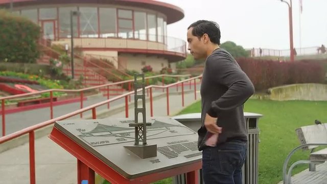 A man looks at a display about the golden gate bridge in a park and then takes a picture
