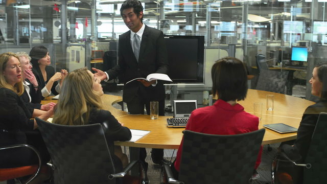 An attractive ethnically ambiguous young businessman wearing a formal suit gives a presentation to several people in a glass-walled office
