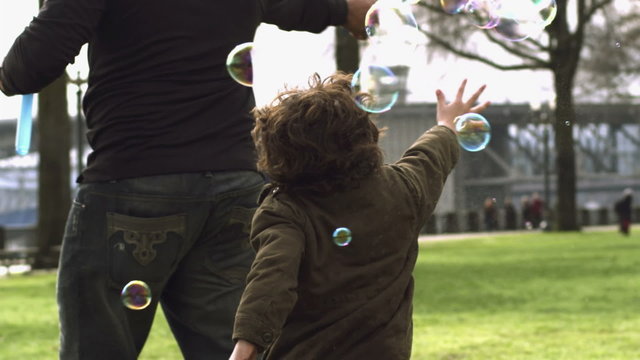 Two young boys chase bubbles blown by their farther around a park