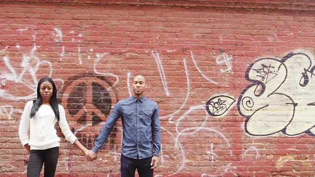 A man and woman stand in front of brick wall with graffiti holding hands, and look into the camera, then turn and stare at each other