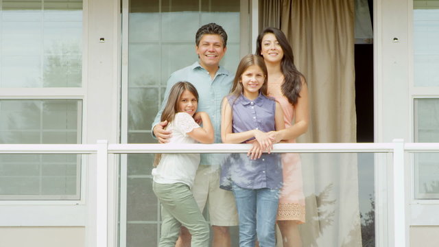 A hispanic family stand on a balcony while smiling and laughing at the camera, during the day