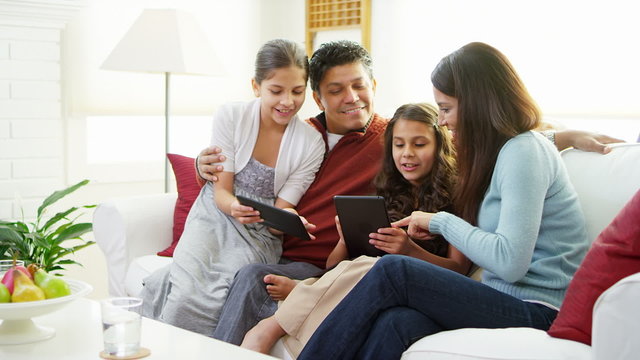 A family of four sit on the couch and play with tablets then smile