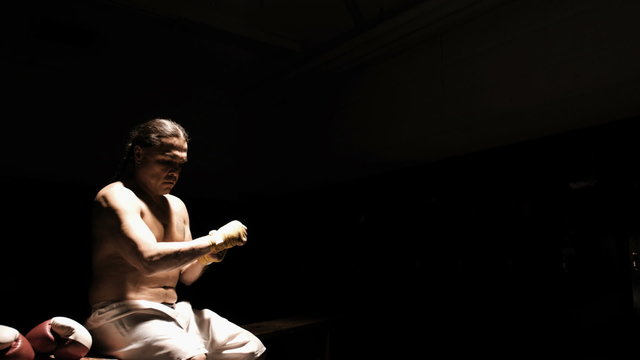 An older boxer wrapping his hands before a fight on a bench in the dark