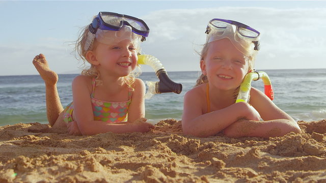 Two sisters lay in the sand together and smile while wearing their snorkel gear