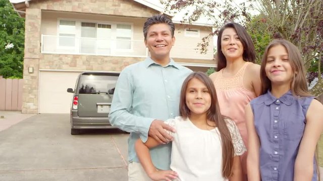 A mother, father and two daughters stand in their driveway and smile and laugh at the camera
