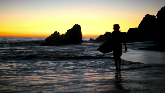 Silhouetted surfer walks along a beach looking at the waves at sunset in Mexico 