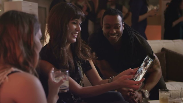 Group of young adults at a house party look at photos of themselves on a tablet