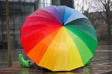 Eine Frau sitzt mit großem bunten Regenschirm und Gummistiefeln auf einer Bank in der Stadt