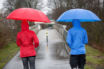 Ein Paar geht mit Regenkleidung und Schirm auf eine Brücke zu. In Rot und Blau