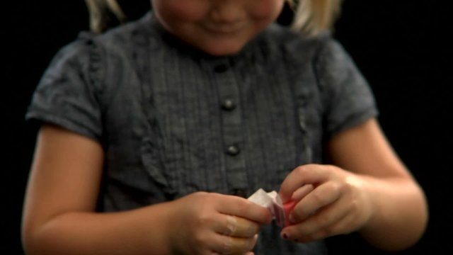A  young blonde girl with pigtails unwraps a piece of bubble gum and chews it