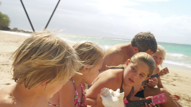 A family sits in the sand at the beach and play ukulele and blow conch shells
