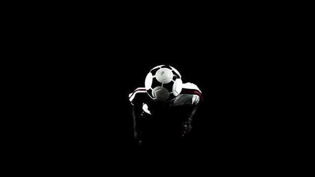Juggling a soccer ball in place on a black floor in the shadows. 